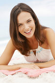 A brunette woman lying on a pink shawl by the sea wearing a white summer dress