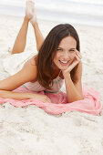 A brunette woman lying on a pink shawl by the sea wearing a white summer dress