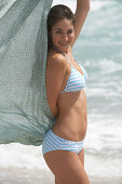 Junge, dunkelhaarige Frau in blau-weiß gestreiftem Bikini mit Tuch am Strand