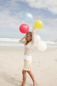 A blond woman on a beach wearing a light cardigan and shorts and holding balloons
