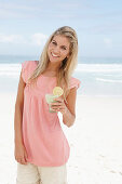 A young woman on a beach with a smoothie wearing a pink top and shorts