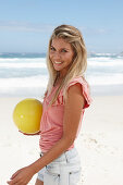 A young woman on a beach with a yellow ball wearing a pink top and denim shorts