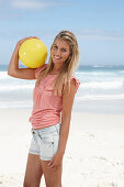 A young woman on a beach with a yellow ball wearing a pink top and denim shorts