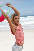 A young woman on a beach with balloons wearing a pink top and denim shorts