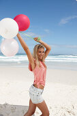 Junge Frau mit Luftballons im rosa Top und Jeansshorts am Strand