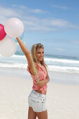 A young woman on a beach with balloons wearing a pink top and denim shorts