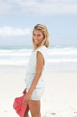 A blonde woman on the beach wearing a light t-shirt and denim shorts holding a red hat