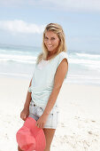 A blonde woman on the beach wearing a light t-shirt and denim shorts holding a red hat