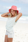 Blonde Frau mit rotem Hut in hellem T-Shirt und Jeansshorts am Strand