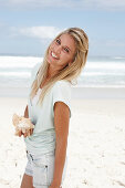 A blonde woman on the beach wearing a light t-shirt and holding a shell