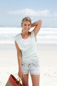 A blonde woman on the beach wearing a light t-shirt and denim shorts holding a wicker bag