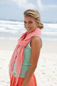 A blonde woman on a beach wearing a scarf, a turquoise top and a salmon pink skirt