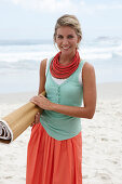 A blonde woman on a beach wearing a turquoise top, a red necklace and a salmon-pink skirt