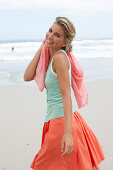 A blonde woman on a beach with a scarf wearing a turquoise top and a salmon-pink skirt