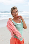 A blonde woman on a beach with a scarf wearing a turquoise top and a salmon-pink skirt