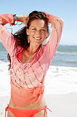 A brunette woman on a beach wearing a pink sequinned blouse and a bikini