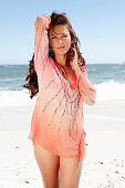 A brunette woman on a beach wearing a pink sequinned blouse and a bikini