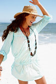 A brunette woman wearing a hat, a necklace and a light-blue beach dress by the sea