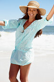 A brunette woman wearing a hat and a light-blue beach dress by the sea