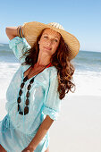 A brunette woman wearing a hat, a necklace and a light-blue beach dress by the sea