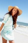 Brünette Frau mit Hut und Halskette in hellblauem Strandkleid am Meer