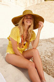 A blonde woman on a beach wearing a hat, a yellow knitted top and a bikini