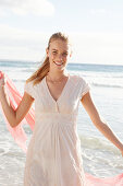 A blonde woman by the sea with a salmon-pink scarf and wearing a white dress