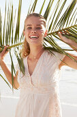 A blonde woman by the sea holding a palm frond wearing a white dress