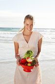 A blonde woman by the sea with a bowl of fruit and vegetables wearing a white dress