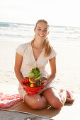 A blonde woman by the sea with a bowl of fruit and vegetables wearing a white dress