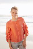 A young blonde woman wearing an orange blouse and light trousers by the sea