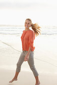 A young blonde woman wearing an orange blouse and light trousers by the sea