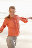 A young blonde woman wearing an orange blouse and light trousers by the sea