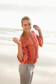 Junge blonde Frau in orangefarbener Bluse und heller Hose am Meer