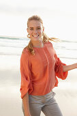 A young blonde woman wearing an orange blouse and light trousers by the sea