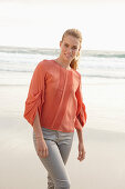 A young blonde woman wearing an orange blouse and light trousers by the sea