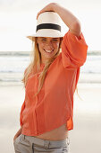 Junge blonde Frau mit Hut in orangefarbener Bluse und heller Hose am Meer