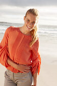 Junge blonde Frau in orangefarbener Bluse und heller Hose am Meer