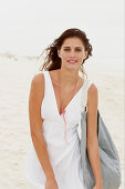 A brunette woman by the sea with a bag wearing a white summer dress