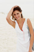 A brunette woman by the sea with a bag wearing a white summer dress