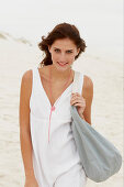 A brunette woman by the sea with a bag wearing a white summer dress