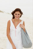 A brunette woman by the sea with a bag wearing a white summer dress