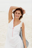 A brunette woman by the sea with a bag wearing a white summer dress