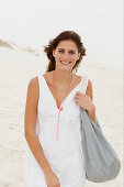 A brunette woman by the sea with a bag wearing a white summer dress