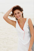 A brunette woman by the sea with a bag wearing a white summer dress