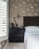 Bed with black headboard and black bedside cabinet against beige floral wallpaper