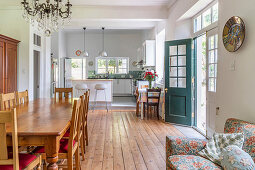 Long wooden dining table and chairs in open-plan interior with wooden floor and kitchen in background