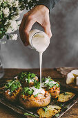 Man's hand pouring sour cream on baked stuffed potatoes with cheese, vegetables and rucola on wooden table