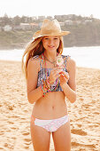 A girl on a beach wearing a hat and a bikini eating an ice cream