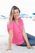 A blonde woman sitting in the sand wearing a pink blouse and jeans